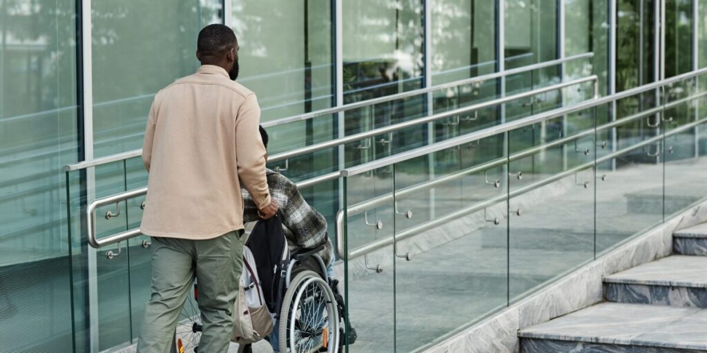 A man is pushing a person sitting in a wheelchair up an accessibility ramp.