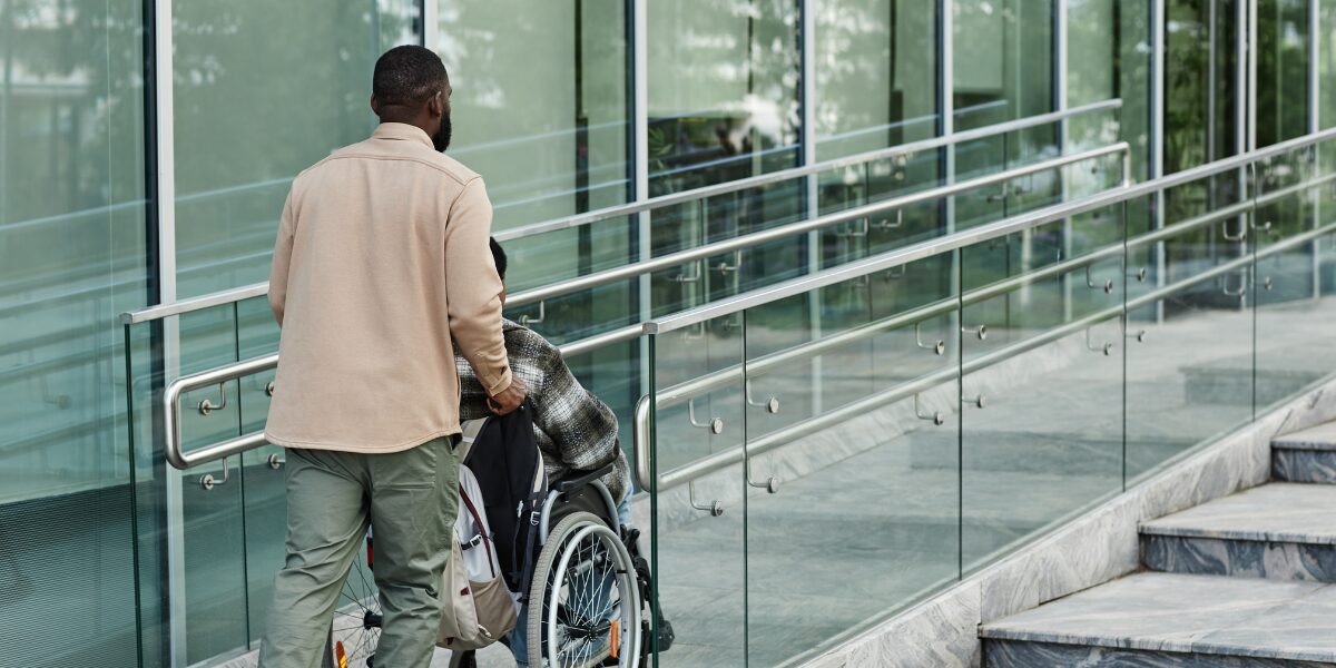 A man is pushing a person sitting in a wheelchair up an accessibility ramp.