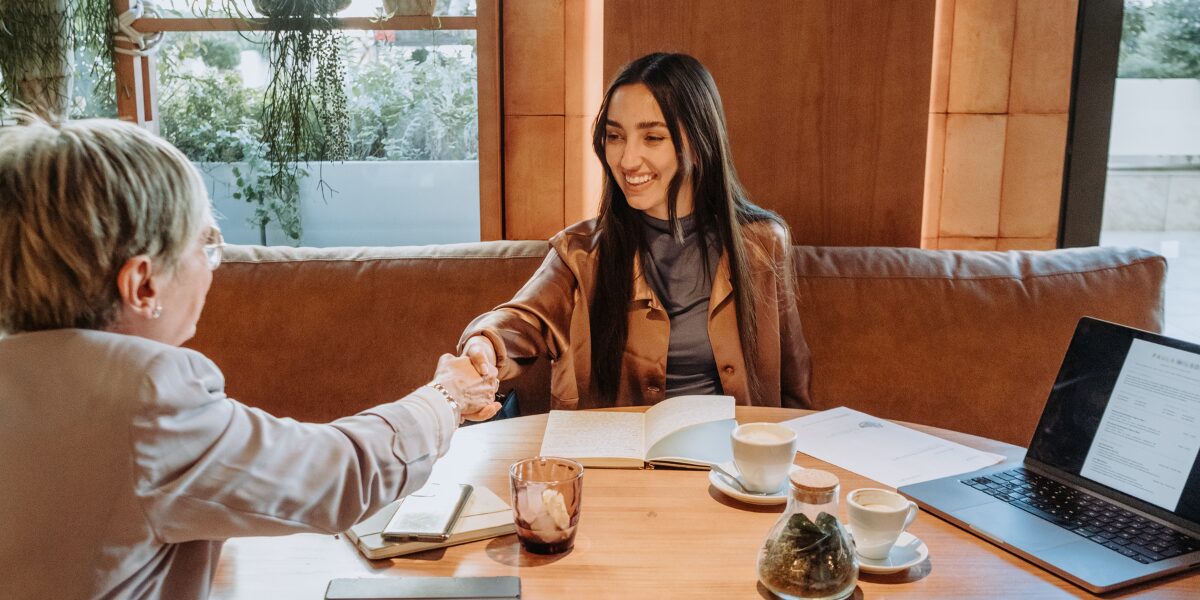 Two women are sat across from one another at a table and they are reaching out to shake one another's hand.