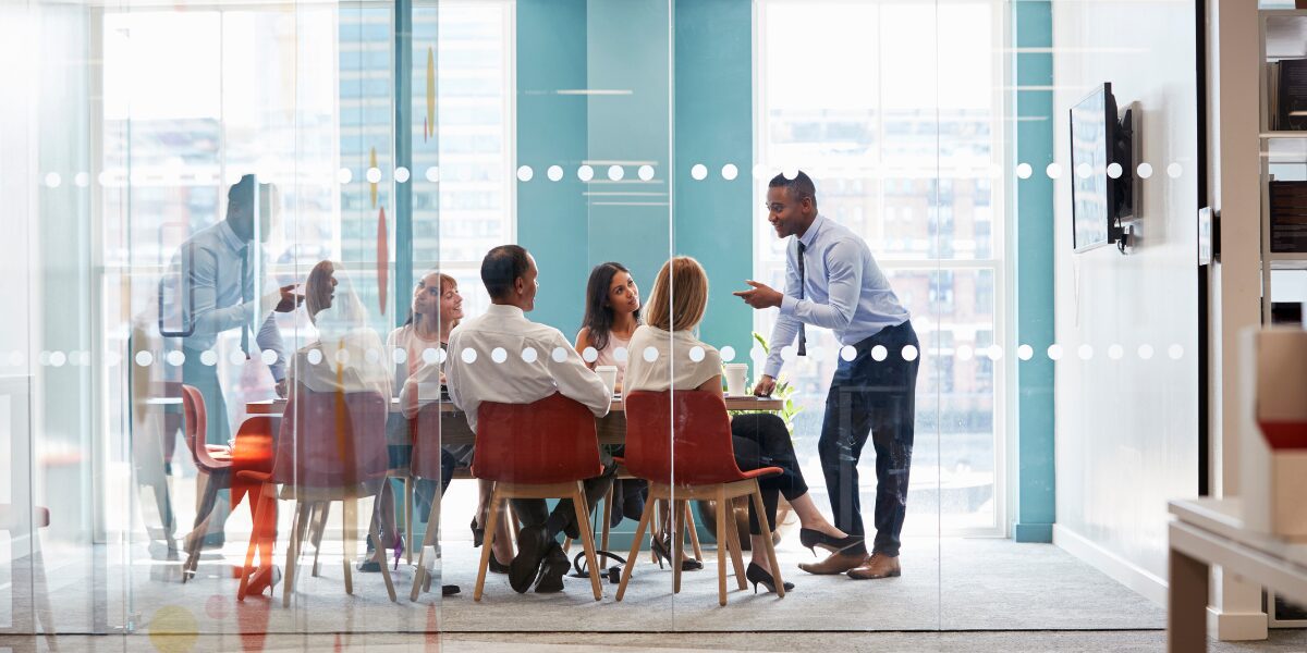 A team of people are sat in a boardroom listening to a leader present.