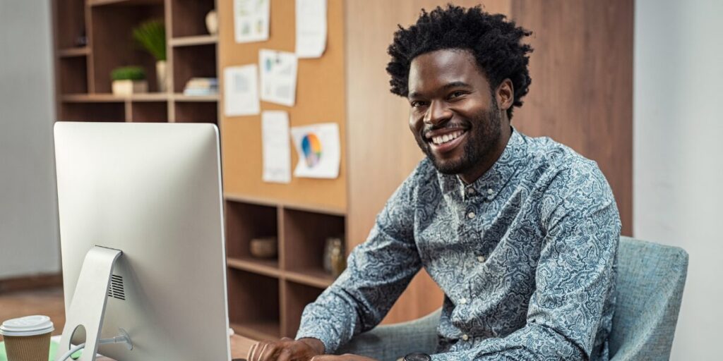 A man is sat at a computer working and smiling at the camera.
