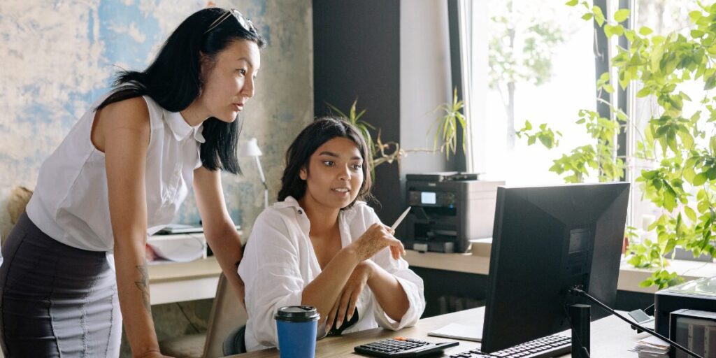Two women are working together and reviewing a webpage on a computer.