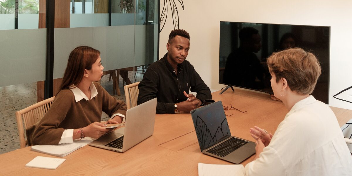 Three people are sat at a boardroom table working and conversing.