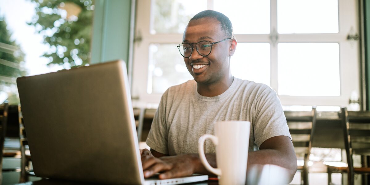A man is sat at a desk working on a laptop and smiling.