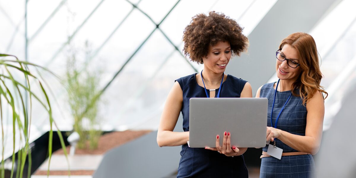 Two women are stood in an office space reviewing a laptop and conversing.